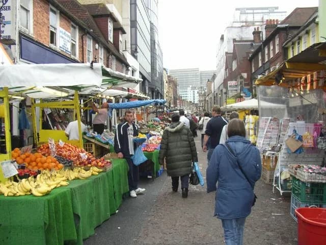 A man in a black and white jacket stands beside a fruit stall on a bustling market street.