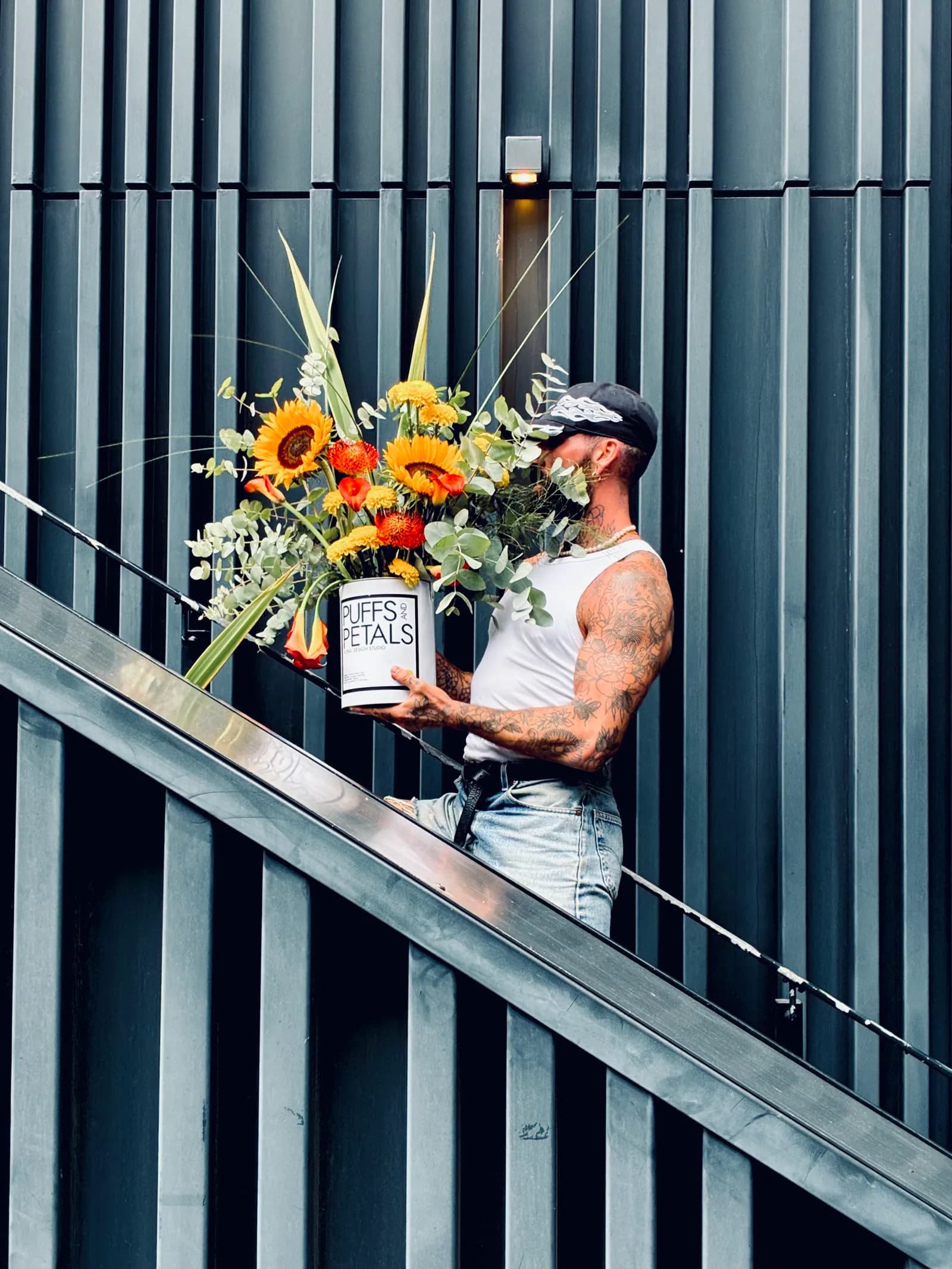 A man with tattoos on his arms, wearing a white tank top and a cap, walks up a dark grey staircase carrying a large bouquet of sunflowers and other flowers in a white box that says "Puffs and Petals."
