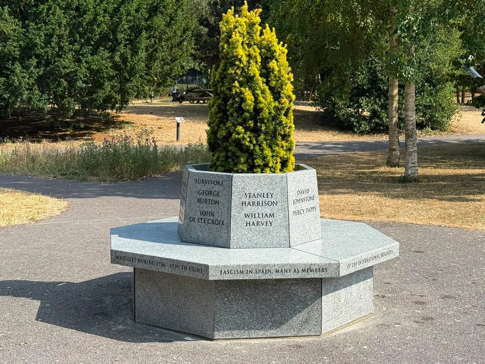 A gray stone memorial with engraved names and text sits on a paved area, flanked by green trees and foliage under a bright sky.