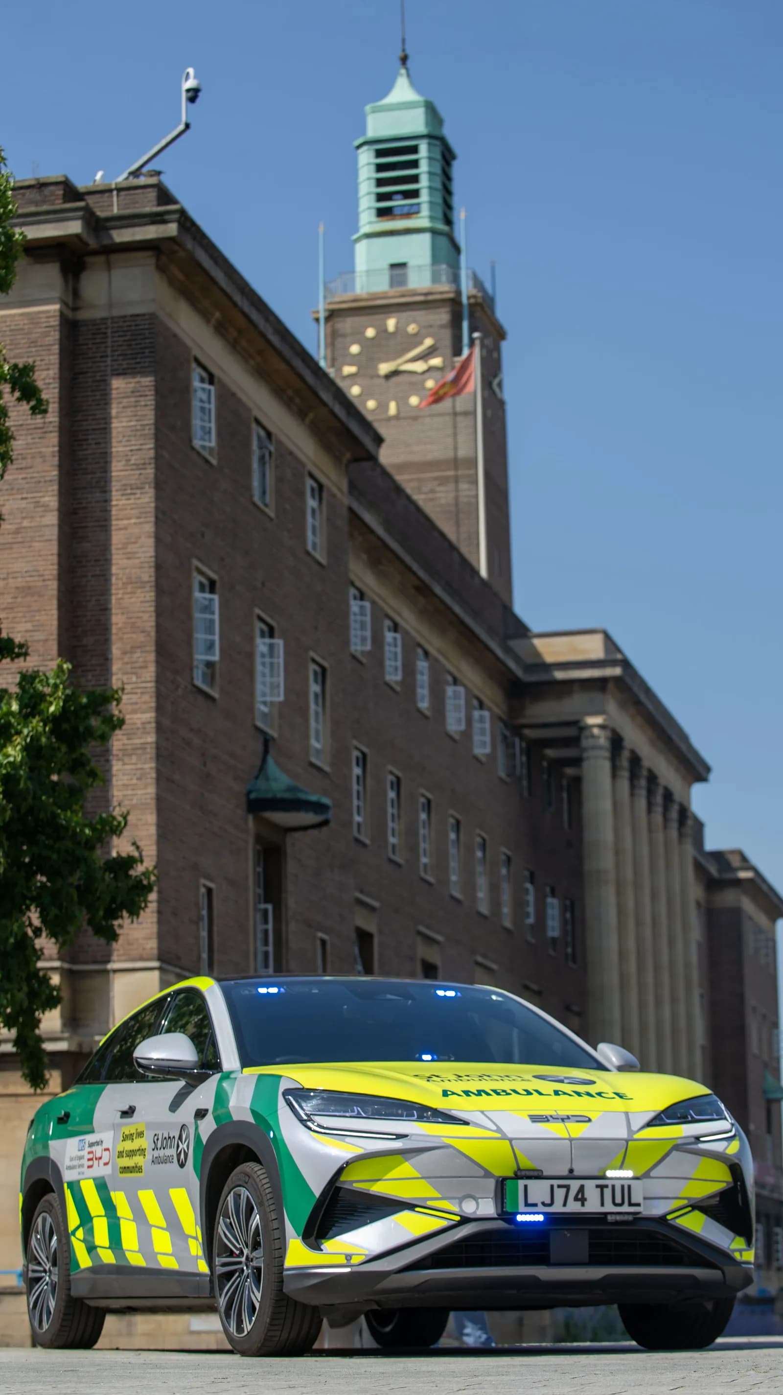 A modern electric ambulance with green and yellow reflective markings is parked in front of a brick building with a large clock tower.