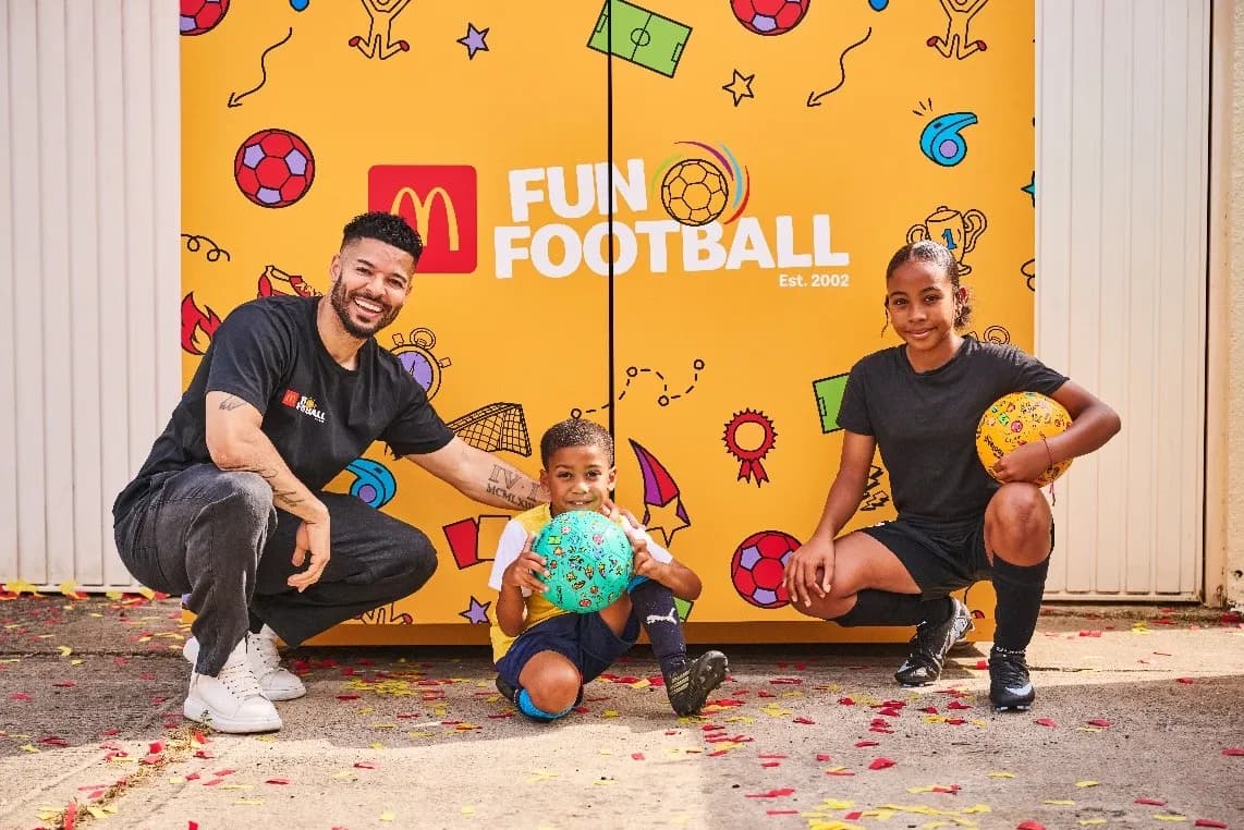 A man, a young boy, and a young girl squatting and smiling in front of a yellow banner promoting Fun Football, with confetti on the ground.