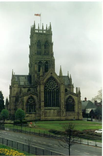 Lichfield Cathedral with a flag flying from its tower, surrounded by green lawns, a road in the foreground, and residential buildings in the background under an overcast sky.