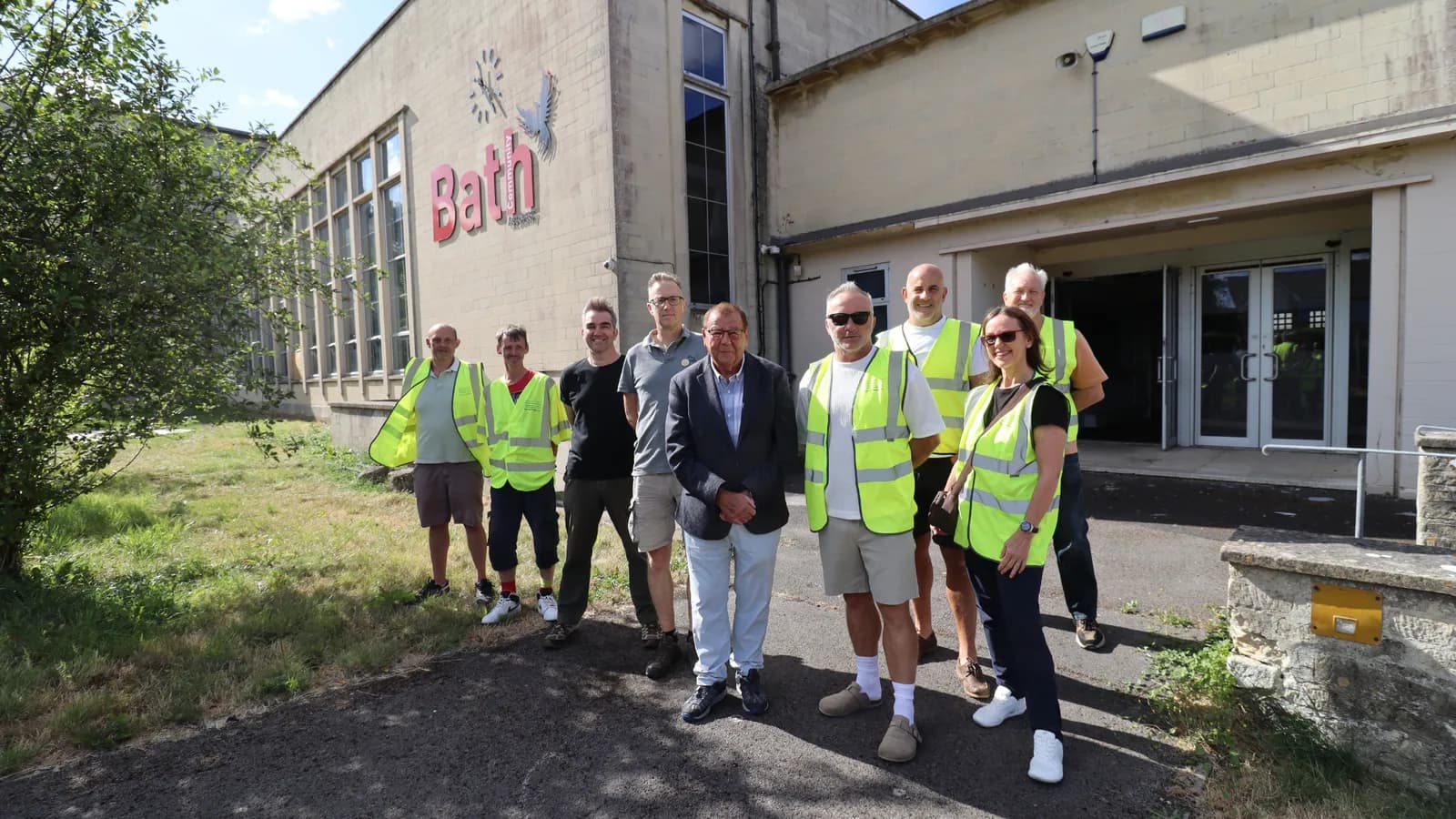 A group of people, some wearing high-visibility vests, stand in front of a building with the word "Bath" and a clock painted on it.