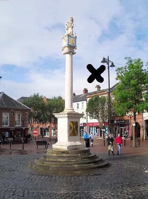 A tall, white stone column topped with a small golden lion statue stands in the center of a cobblestone town square framed by buildings and trees under a cloudy sky.