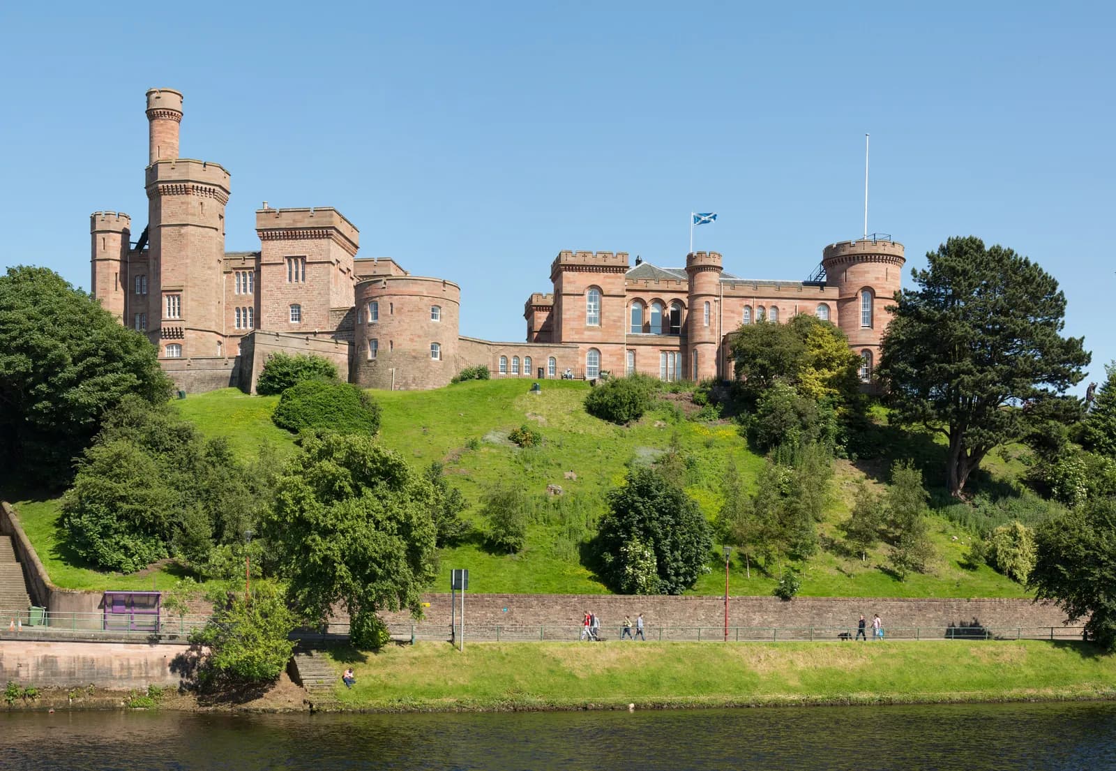 A large, historic brick castle with multiple turrets sits atop a steep, green grassy hill under a clear blue sky, overlooking a wide river with people walking along its bank.