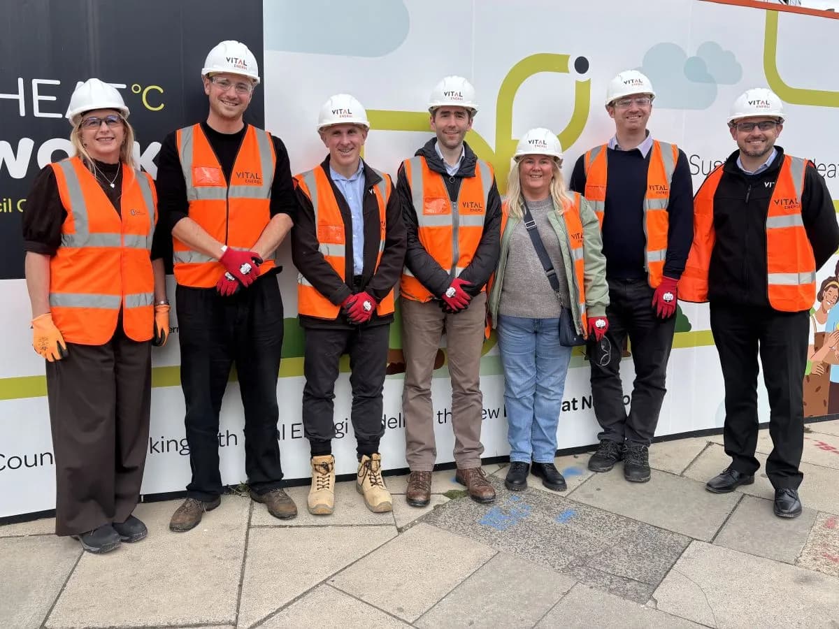 Seven construction workers, four men and three women, wearing orange vests and white hard hats, are standing side by side in front of a white construction hoarding with text and graphics.