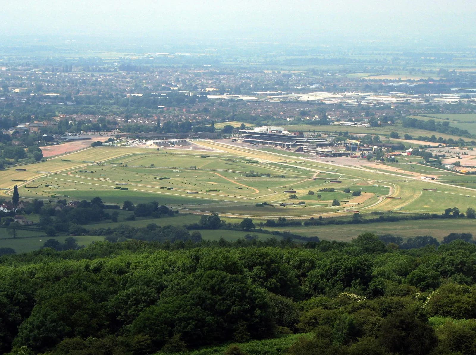 An aerial view captures the sprawling green grounds of a horse racecourse with grandstands and buildings, backed by a distant town and hazy sky.