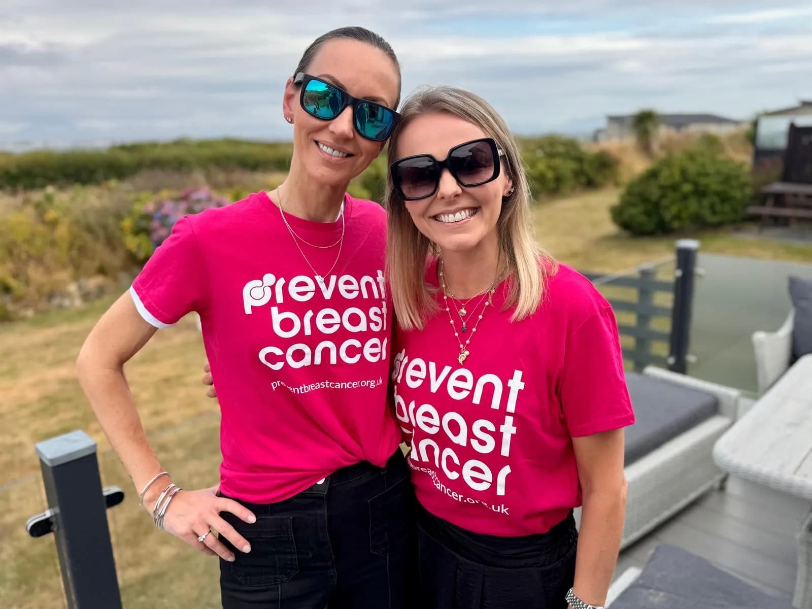 Two women wearing pink “Prevent Breast Cancer” t-shirts and sunglasses smile at the camera, standing outdoors with a blurred background of greenery and outdoor furniture.