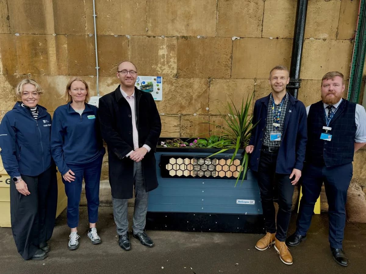 Five people stand next to a large blue planter box with green plants and flowers against a light brown brick wall.