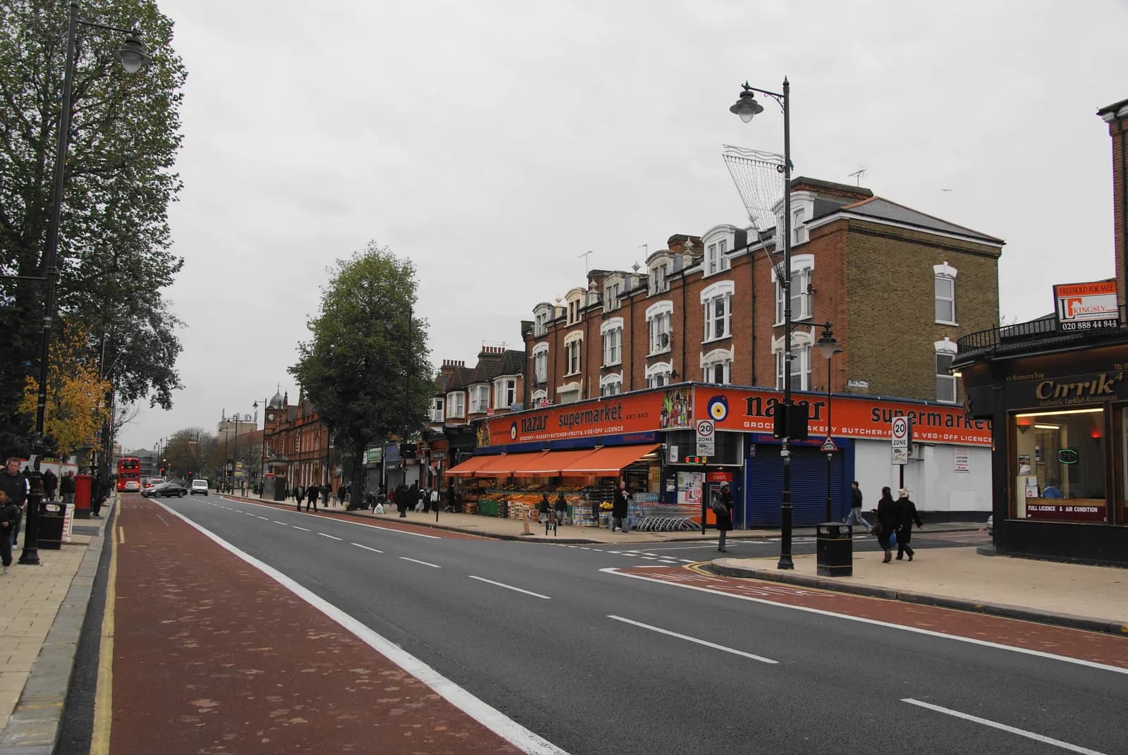 A long, straight road with cars and pedestrians on either side, lined with shops and buildings, under an overcast sky.