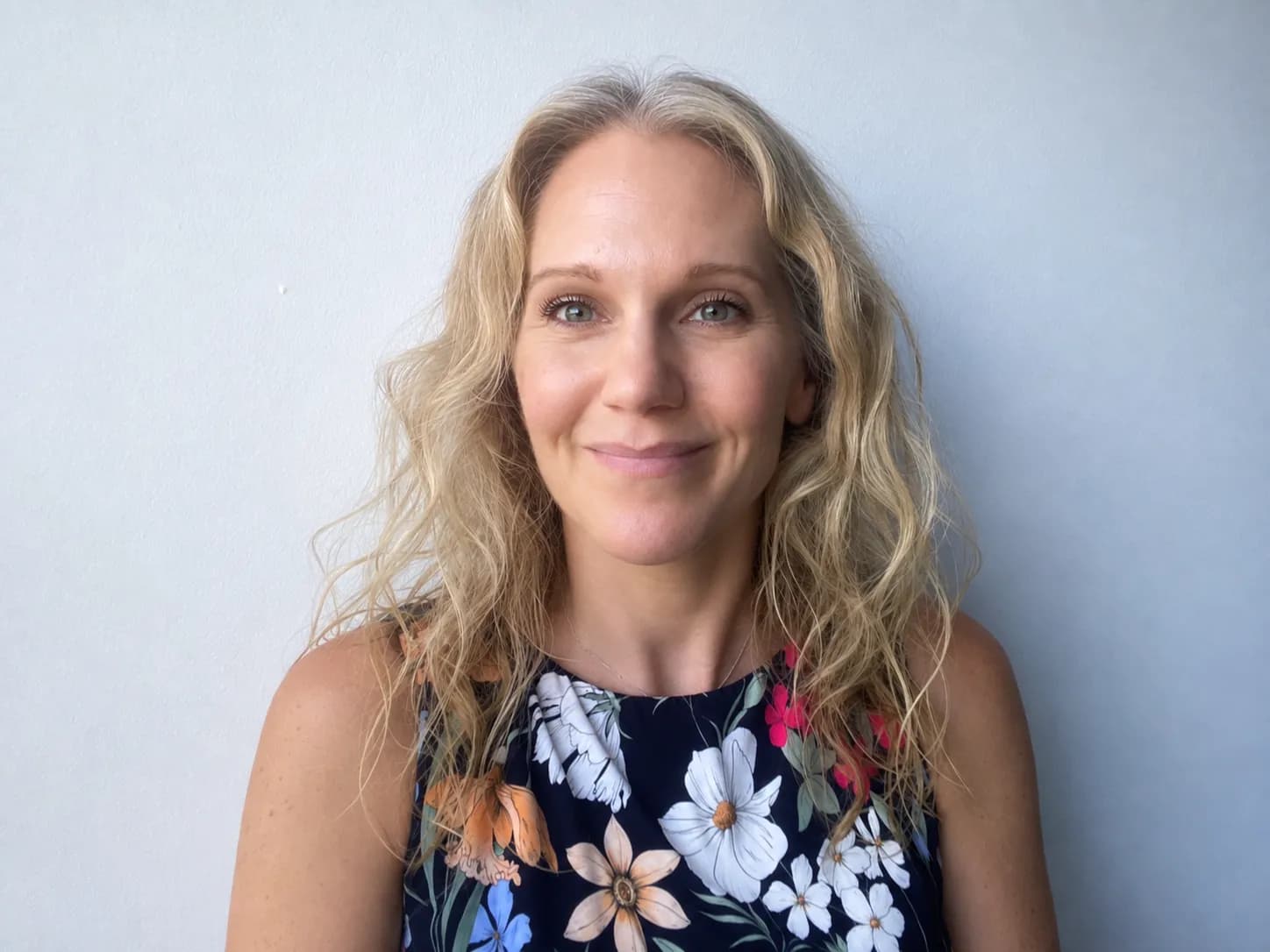 A smiling woman with long wavy blonde hair and a floral top looks directly at the viewer against a plain light background.