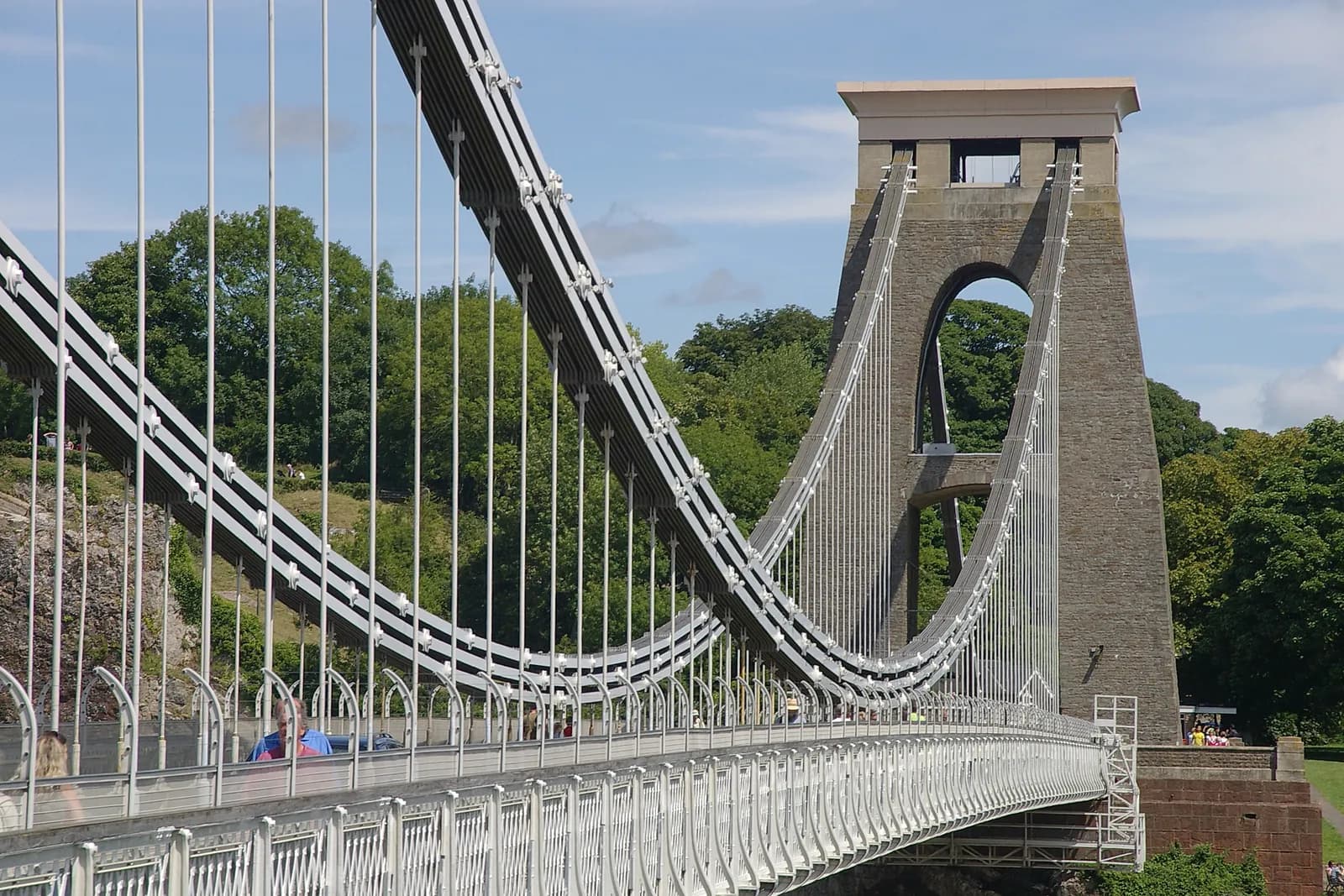 The Clifton Suspension Bridge shows its intricate gray cable structure leading to a large stone tower on a bright sunny day with green trees and a blue sky in the background.
