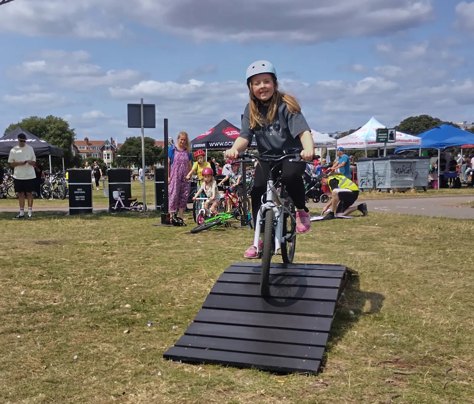 A smiling young girl in a helmet and t-shirt rides a bicycle up a small wooden ramp in a grassy park.