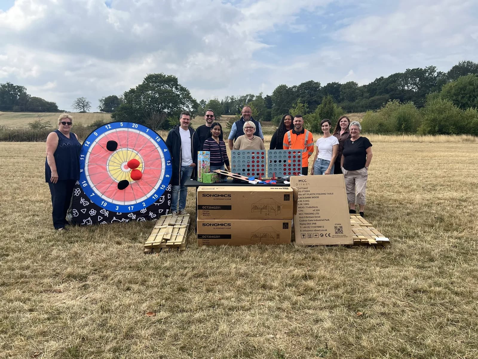 A group of people stand in a grassy field with a large dartboard, giant Connect Four game, and several cardboard boxes of outdoor games.
