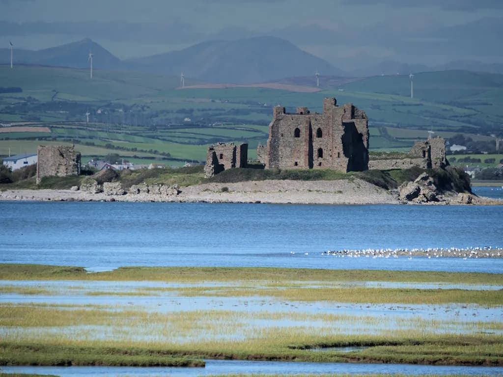 A medieval stone castle in ruins sits on a rocky bluff beside a large body of shimmering blue water, with green fields and distant mountains visible in the background under a blue sky.