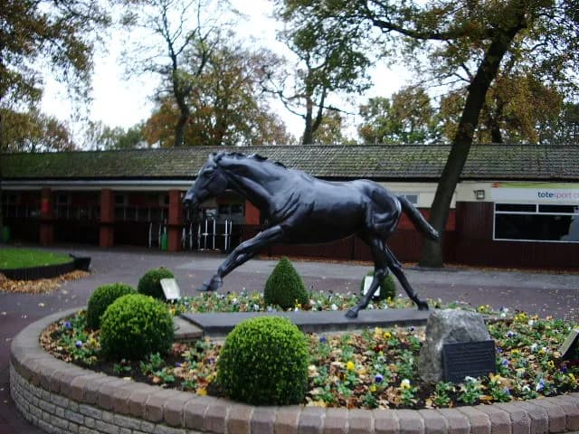 A dark bronze statue of a galloping horse is surrounded by a low brick planter filled with small green bushes and colorful flowers, with a building and trees in the background.