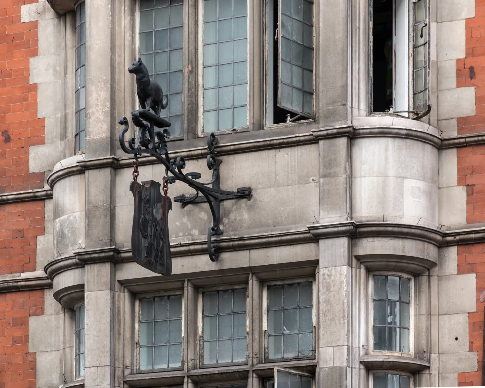 A black wrought iron sign featuring a dog statue on top and a hanging rectangular plaque, attached to a stone building with multiple windows and red brick accents.