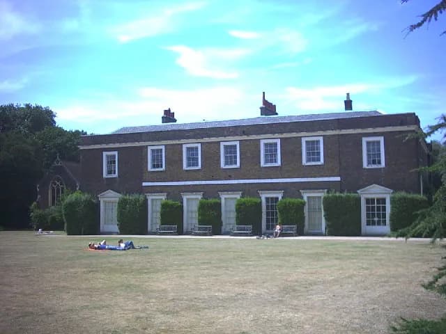 A long, two-story brick building with white-framed windows and doors is seen across a large, dry grass lawn with a few people resting on it under a bright blue sky.