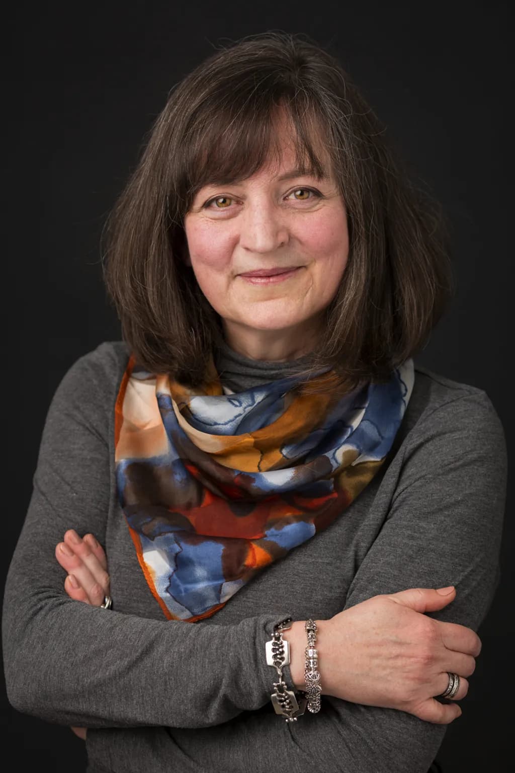 A smiling woman with brown hair and a colorful scarf looks directly at the viewer, her arms crossed in front of her.