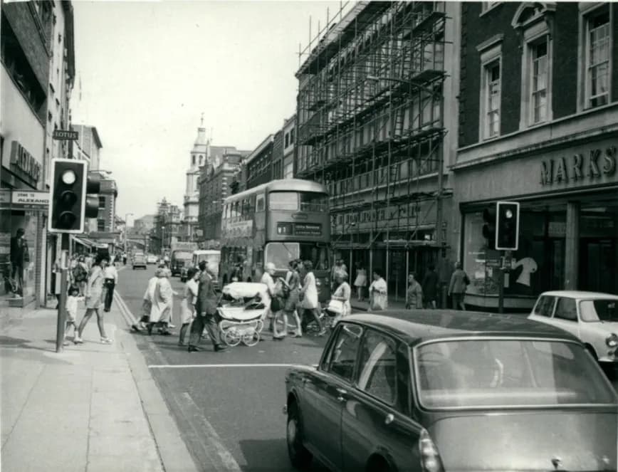 A bustling street scene from the 1960s with pedestrians crossing the road, a double-decker bus, cars, and buildings with scaffolding.