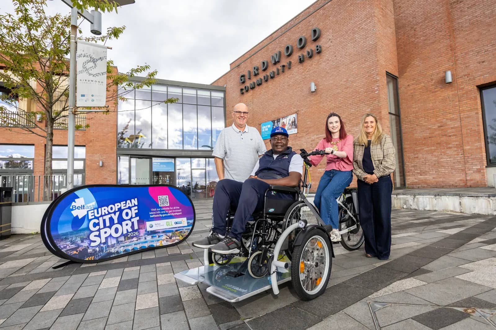 Four people, including one in a wheelchair and one on a bicycle, stand outside a brick building next to a banner advertising Belfast European City of Sport 2026.