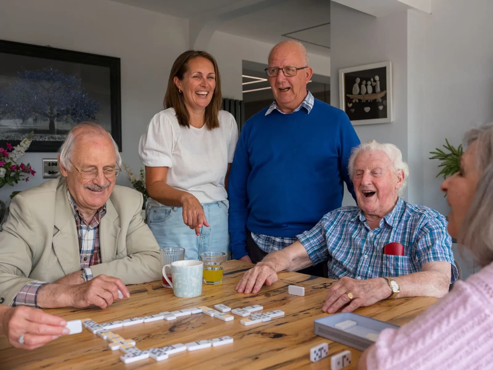 Four elderly people and one younger woman laugh while playing dominoes around a wooden table.