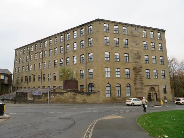 A large, multi-story brick building with many windows sits next to a road, with a few cars and people passing by.