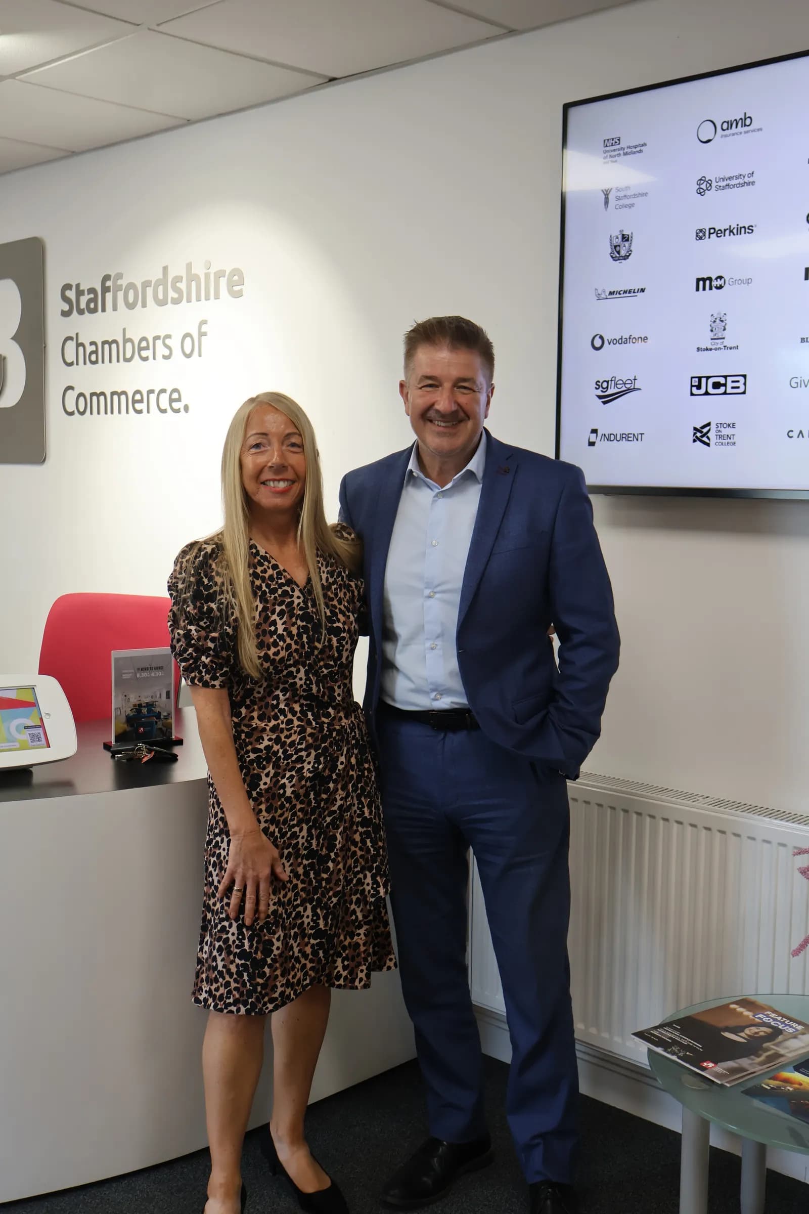 A woman in a leopard print dress and a man in a blue suit stand smiling in front of a white wall with the words "Staffordshire Chambers of Commerce" and several company logos.