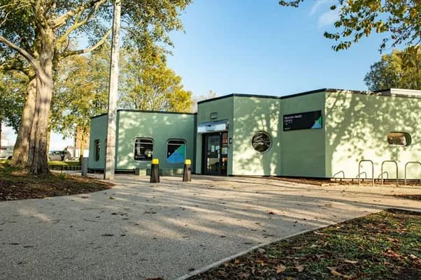 Exterior view of a modern, light green building with large windows and a dark entrance, surrounded by trees and a paved area with bike racks.
