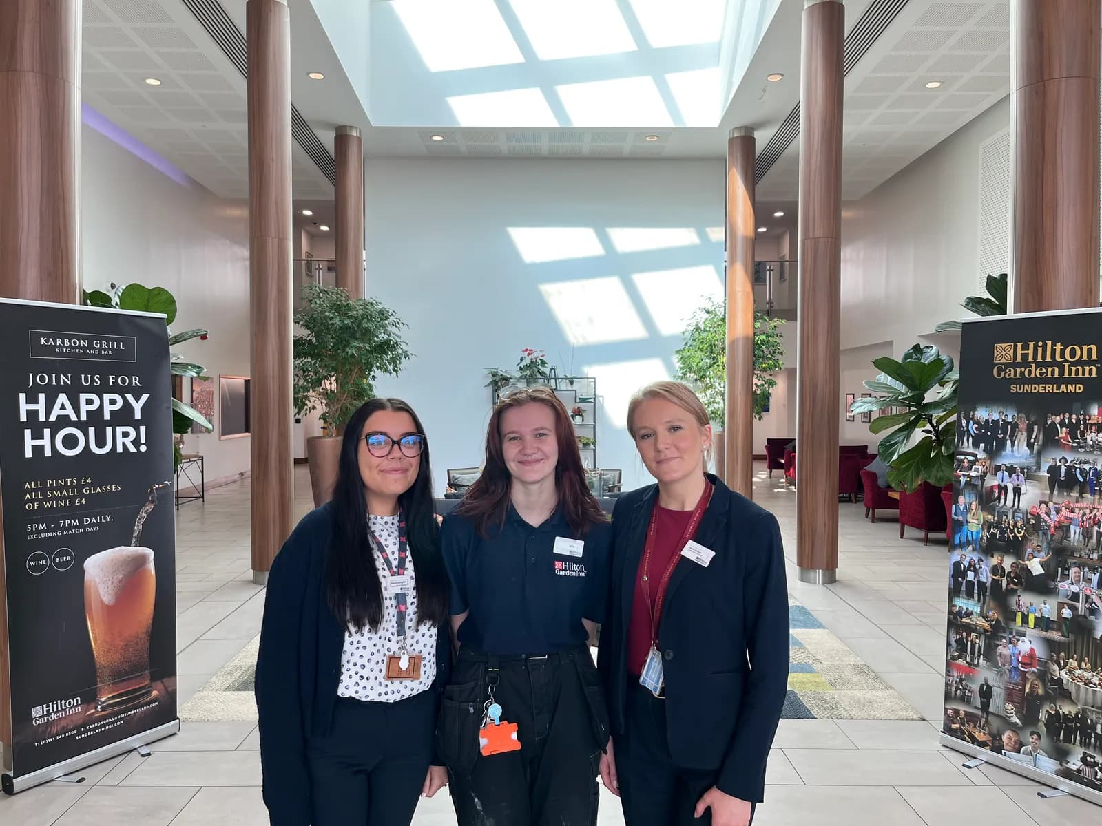 Three women stand side-by-side in a large, modern hotel lobby with high ceilings and large pillars.