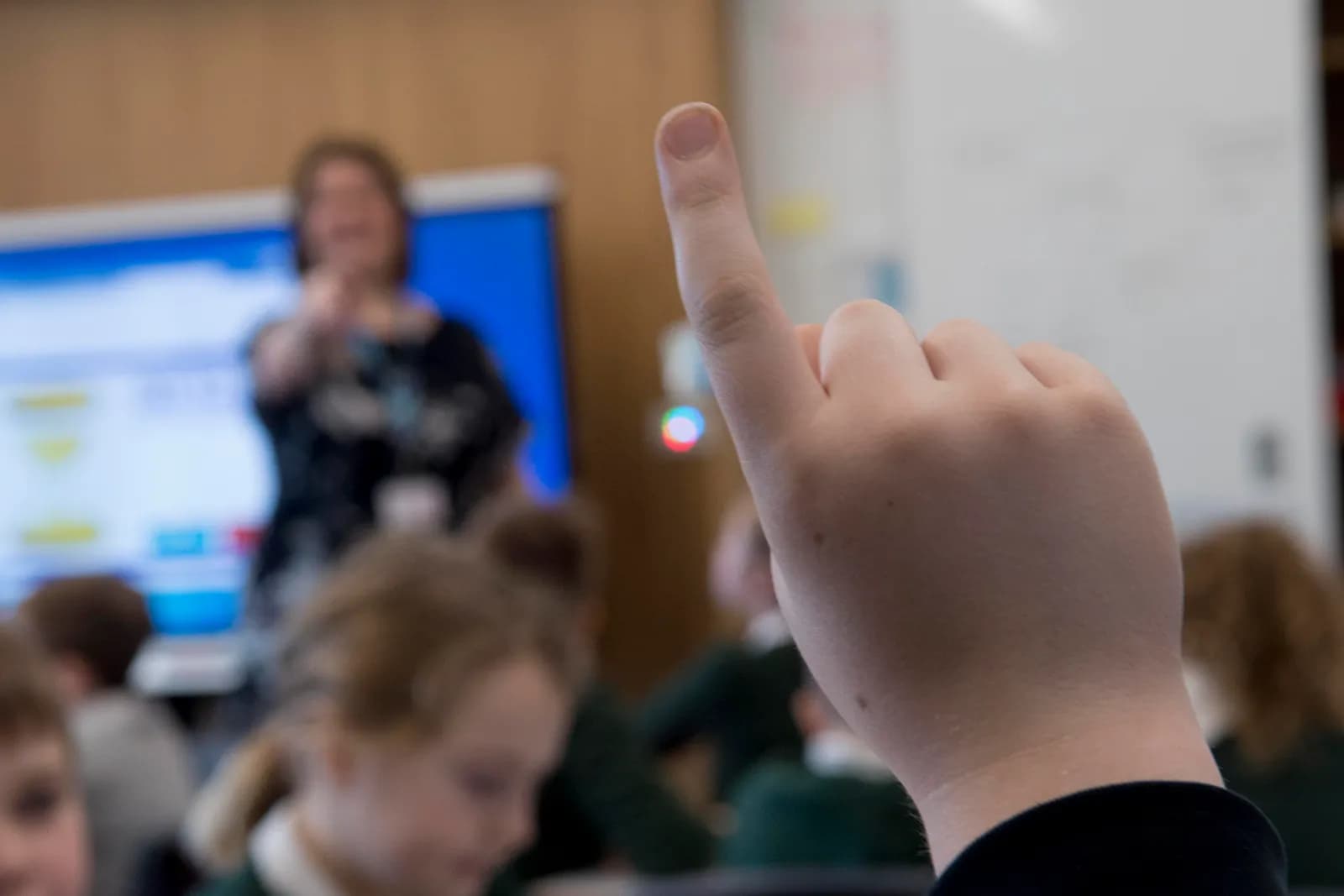 A student's hand is raised in the foreground, with a teacher standing in front of a screen in the blurred background.