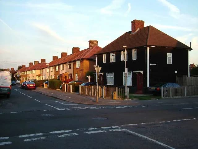 A row of houses, starting with a large black house, extends along a curving suburban street under a bright sky with wispy clouds.