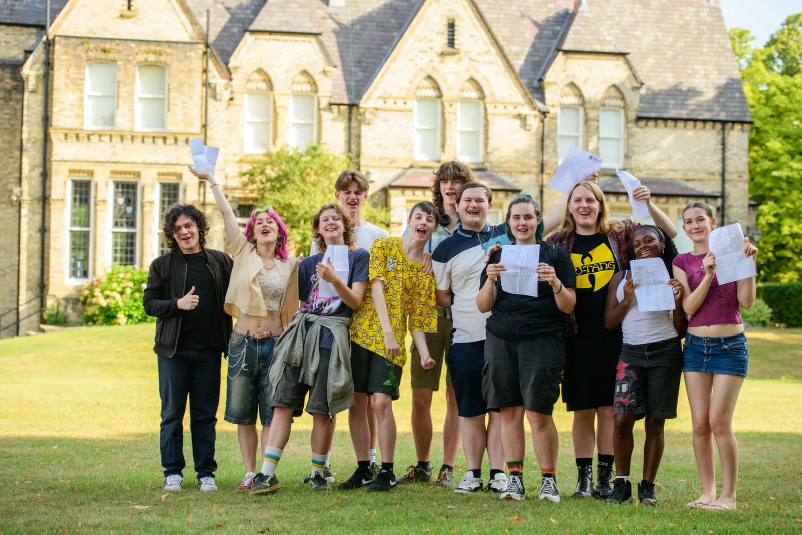 A diverse group of smiling teenagers stands on a grassy lawn in front of a large, old building, holding up papers that appear to be exam results.