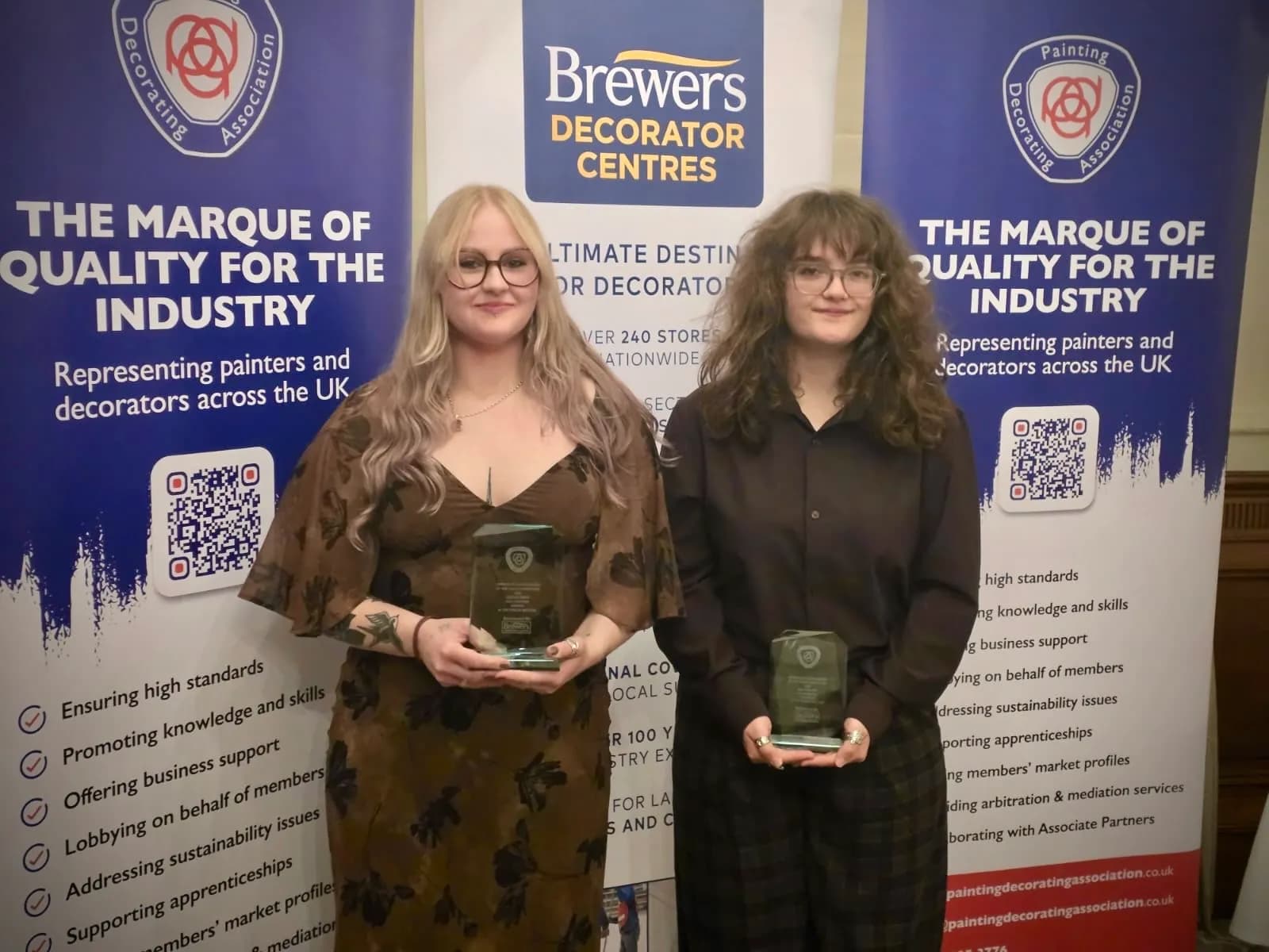 Two young women stand side-by-side holding awards in front of banners with text that reads "The Marque of Quality for the Industry."