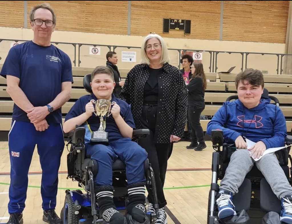 A boy in a wheelchair holding a trophy, flanked by a man and a woman, with another boy in a wheelchair to the right, all smiling in a gymnasium.