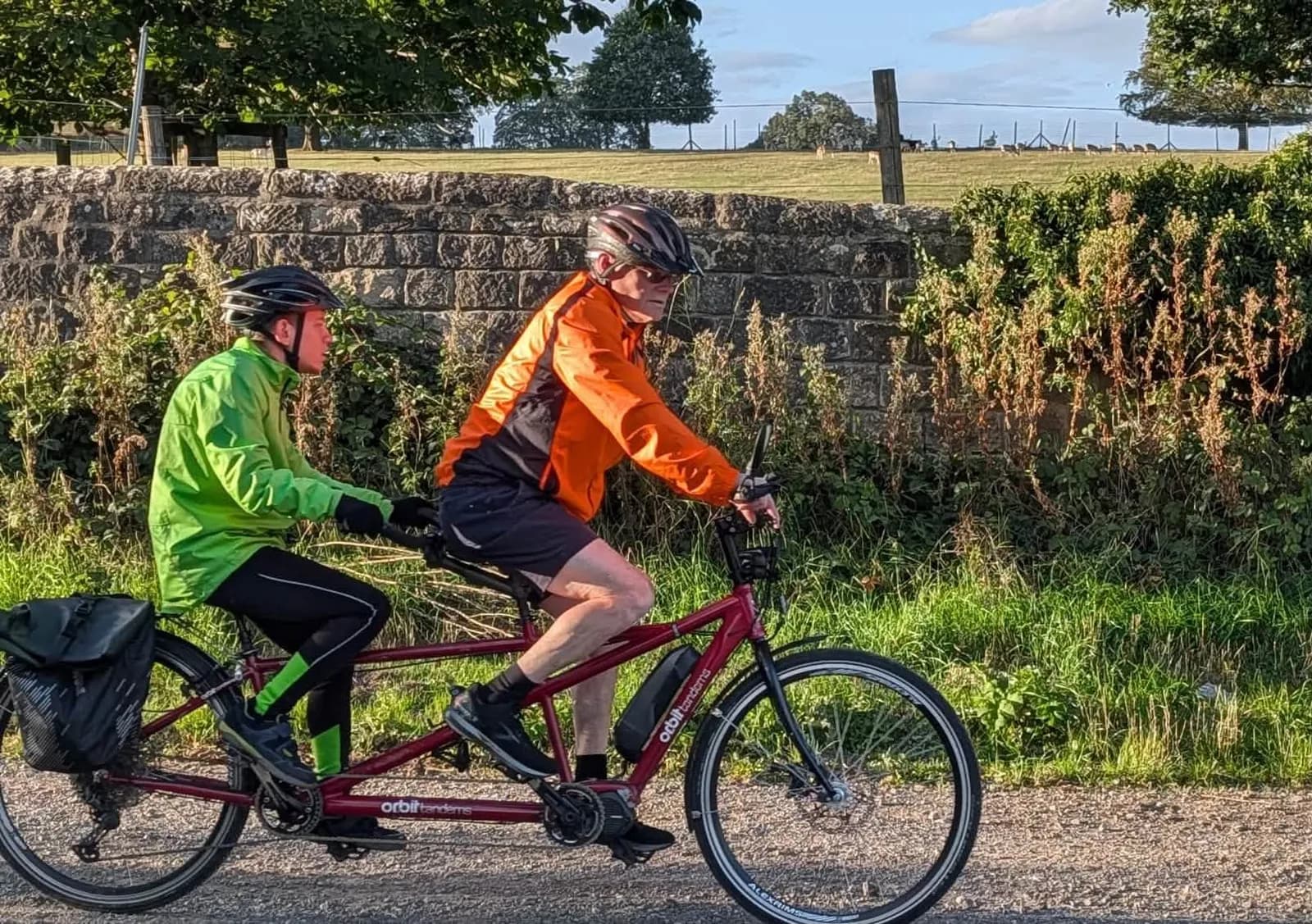 Two people riding a red tandem bicycle on a path alongside a stone wall and green foliage under a partly cloudy sky.