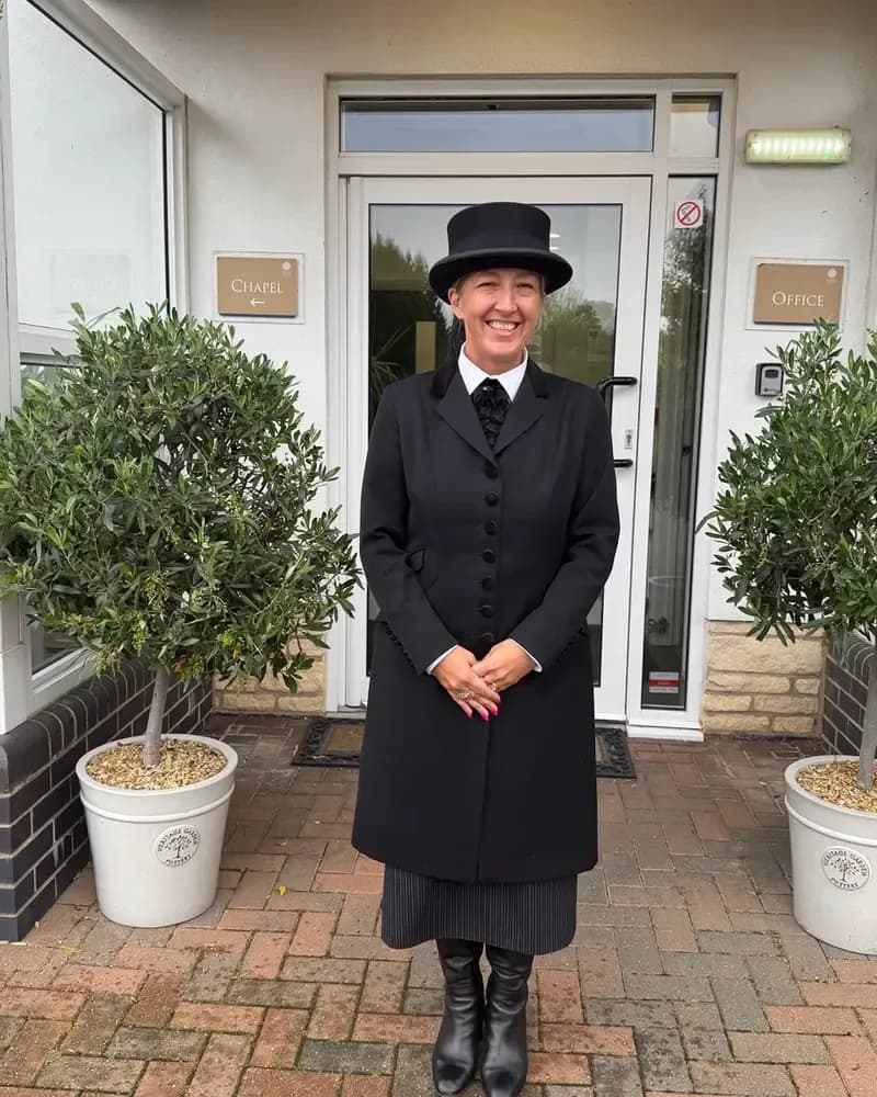 A woman in a black uniform including a top hat stands smiling in front of a white building with signs for "CHAPEL" and "OFFICE".