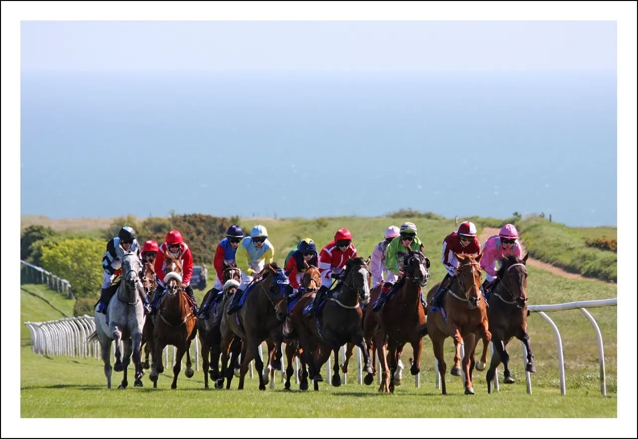 A line of horses and jockeys gallop across a green field with a blue ocean and sky in the background.