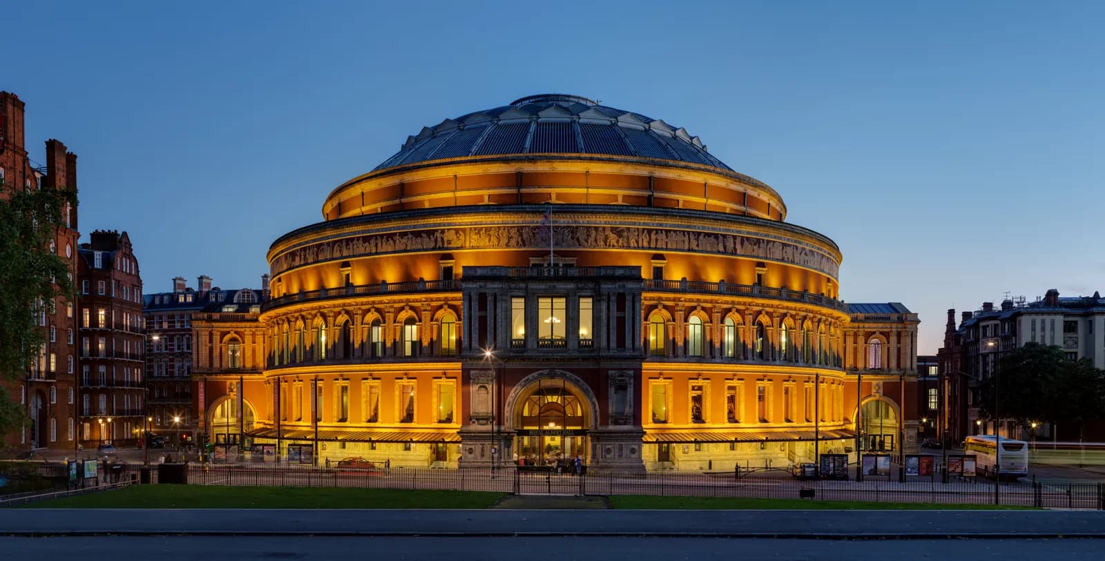 The Royal Albert Hall, a grand, circular building with a large dome, is brightly illuminated at dusk, appearing golden against the deep blue sky.