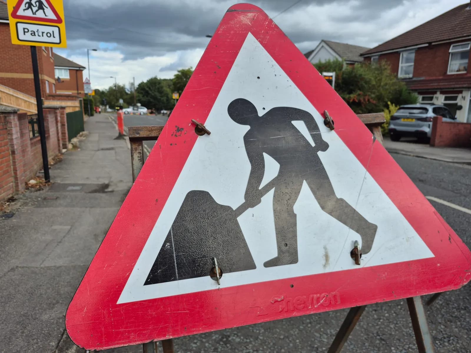 A triangular road sign warns of road works, showing a person digging with a shovel next to a mound of earth, with a residential street visible in the blurred background.