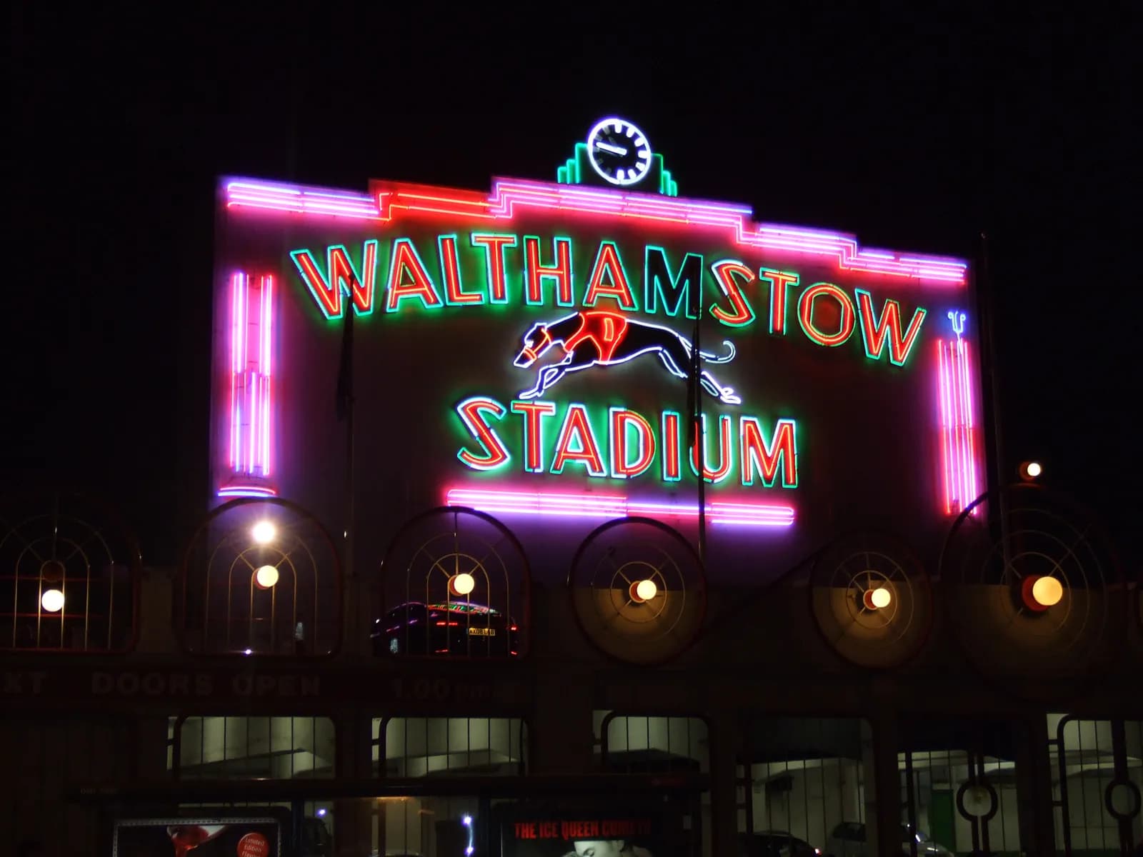 A neon sign for Walthamstow Stadium at night, featuring a green and red text sign, a clock, and a greyhound silhouette.