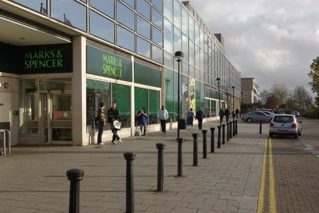 A Marks & Spencer department store with a glass exterior and green signage, with multiple pedestrians walking on the pavement outside and cars parked on the side of the road.