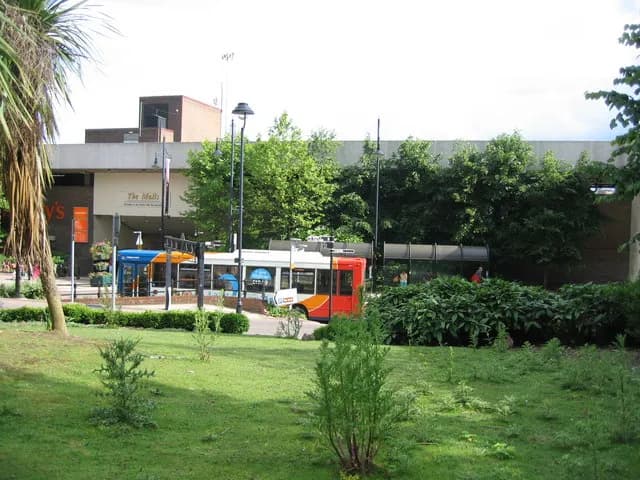 A red and white bus is parked near a building with 'The Malls' written on it, surrounded by green trees and grass on a bright day.