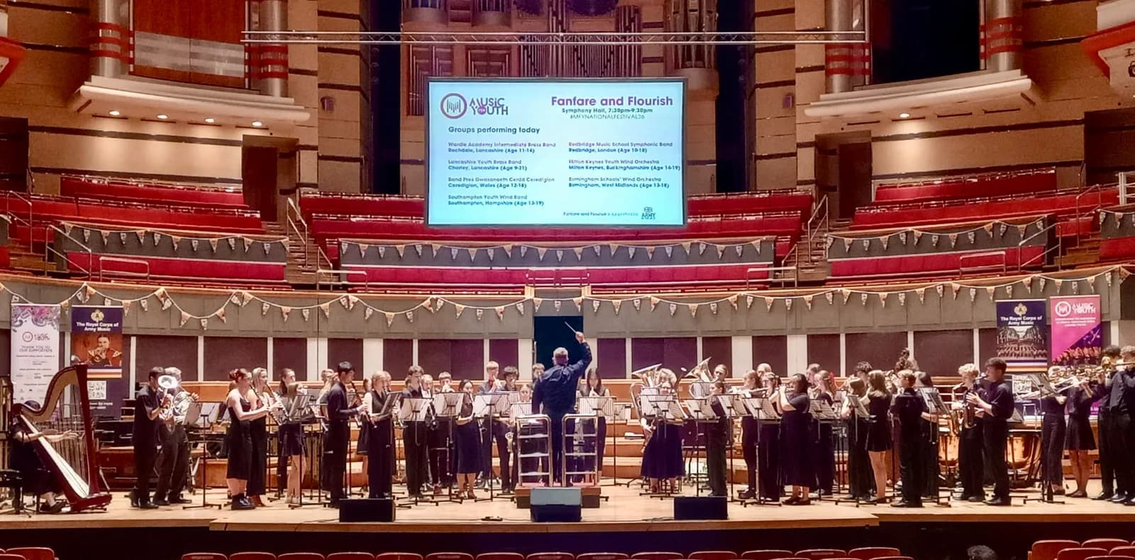 A conductor leads an orchestra of young musicians on a stage in a large auditorium with red seating, under a screen displaying event information.