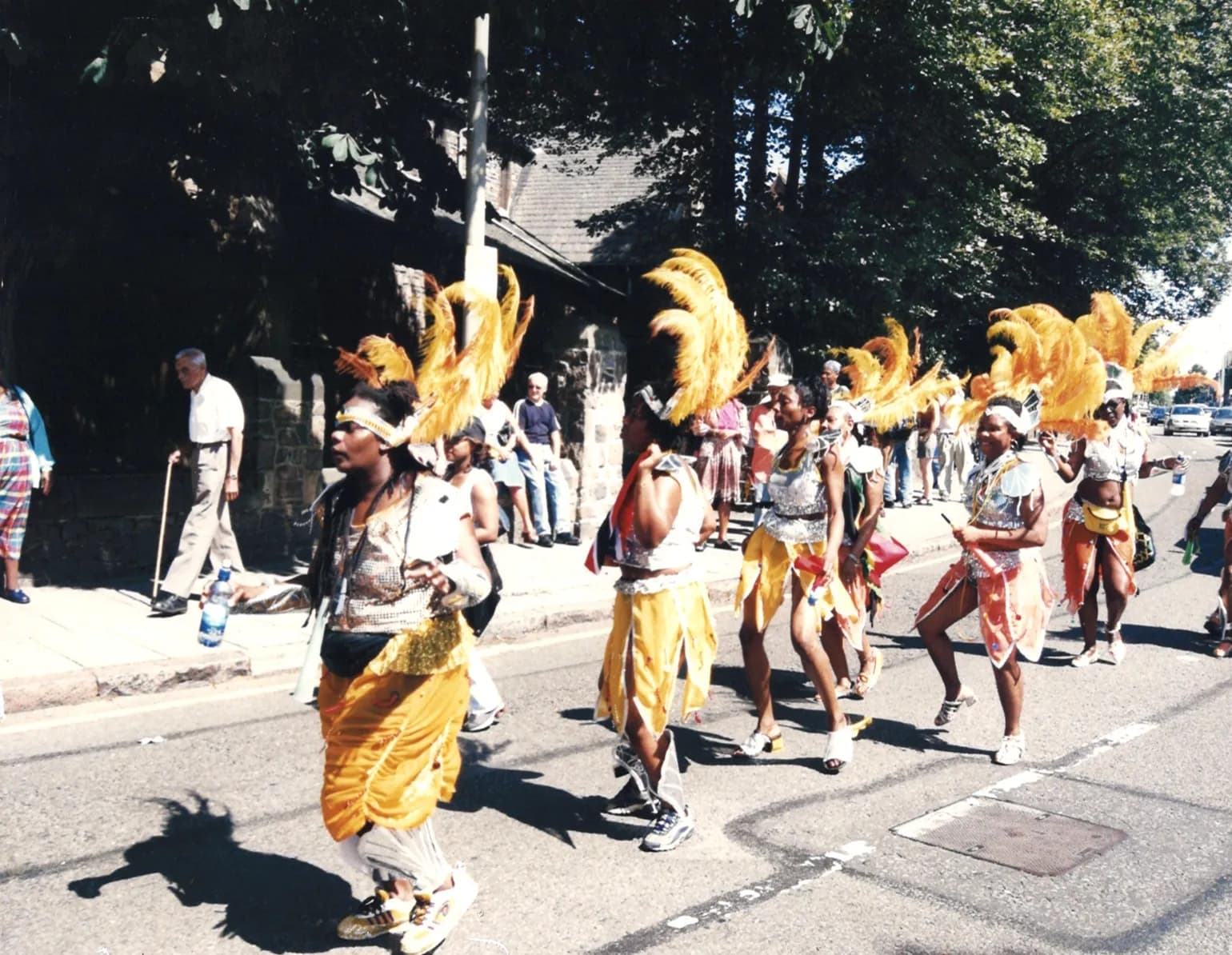 People in carnival costumes with large yellow feather headpieces parade down a sunlit street, with onlookers on the sidewalk and trees in the background.