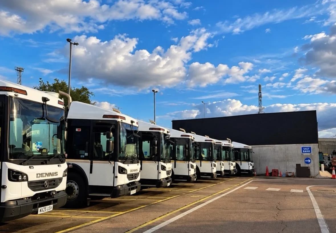 A line of white and black Dennis Dart buses parked in an outdoor lot under a blue sky with white clouds, with a building in the background.