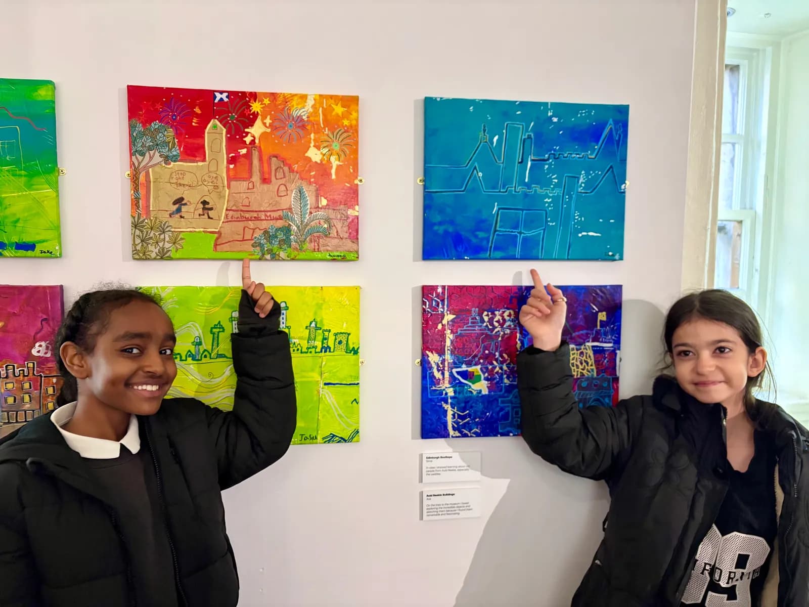 Two smiling girls stand in front of colorful paintings on a wall, each pointing to a different piece of art.