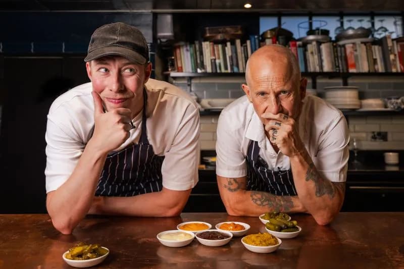 Two chefs, one younger with a cap and one older and bald, lean on a wooden counter with various small bowls of sauces and pickles in front of them.