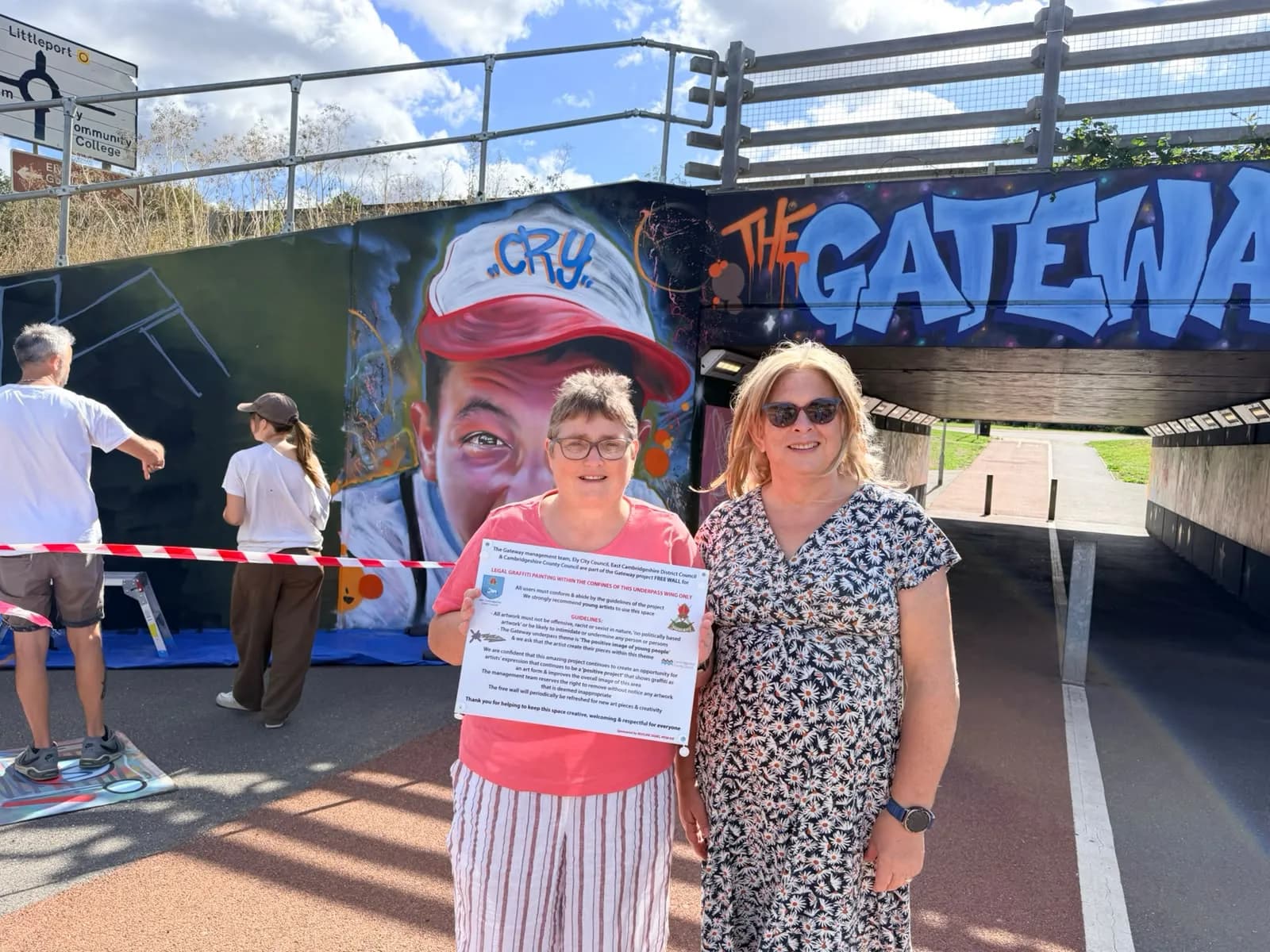 Two women, one holding a sign, stand in front of a graffiti-covered wall that includes a large painted face and the words "THE GATEWAY."