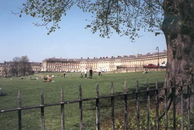 A wide shot of the Royal Crescent in Bath, England, with people relaxing on the green lawn in front of the curved Georgian buildings under a clear sky.